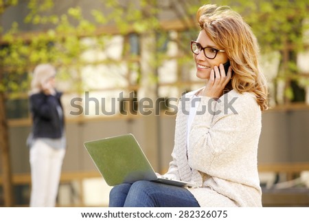 Image of casual businesswoman sitting at outdoor while using her laptop and making call with mobile. Business people at work.