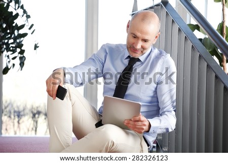 Portrait of senior bank employee sitting at staircase while holding hands digital tablet and handy.