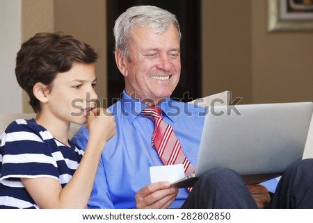 Portrait of teenager boy helping his grandfather how to use credit card for secure online shopping. Grandfather and grandson sitting at sofa and using laptop.