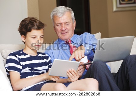 Portrait of grandfather with laptop and his grandson with digital tablet sitting at living room and using credit card for online shopping.