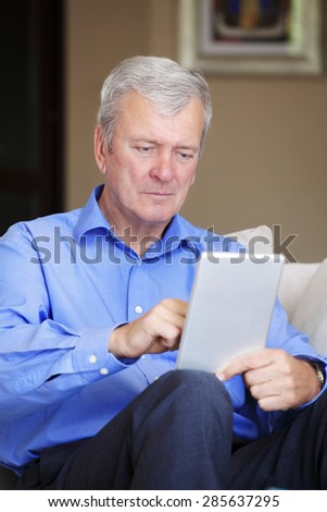 Portrait of senior man sitting at home and holding digital tablet in his hands. Retired man using tablet and touch the screen.