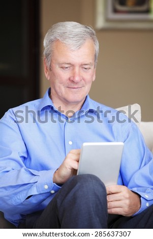 Portrait of senior man sitting at home and holding digital tablet in his hands. Retired man using tablet and touch the screen.