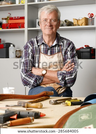 Portrait of proud senior carpenter sitting at his workshop with arms crossed. Small business.