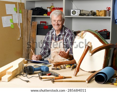 Portrait of senior carpenter holding clipboard and making notes while sitting at his workshop and repair the broken legged chair. Small business at home.