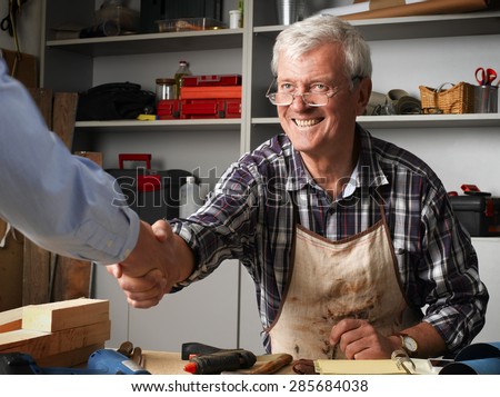 Smiling senior carpenter greeting customer and shaking his hand at workshop. Small business.