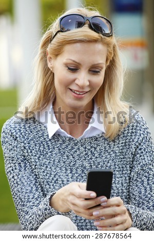 Full length portrait of casual businesswoman sitting at business park while touching her mobile phone. Middle age woman wearing casual clothes