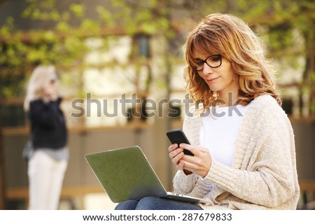 Portrait of busy sales woman sitting at business park and using her laptop while writing text on mobile. Businesswoman working online.