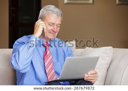 Portrait of active retired man sitting at home and working. Senior businessman using his laptop while making call.