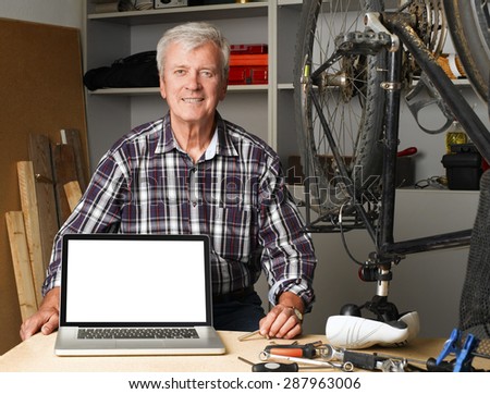 Portrait of senior bike shop owner sitting at desk behind the laptop with white screen. Active old man looking at camera and smiling. Small business.