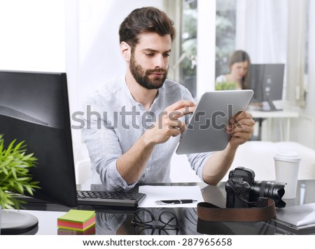 Portrait of young businessman holding digital tablet in his hands while sitting at office desk in front of computer and working. Young professional man touching the screen.