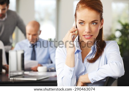 Portrait of attractive business woman sitting at desk and making call while business people working at background. Teamwork at office.
