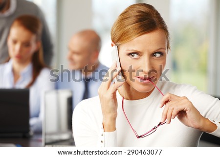 Portrait of attractive business woman sitting at desk and making call while business people working at background. Teamwork at office.