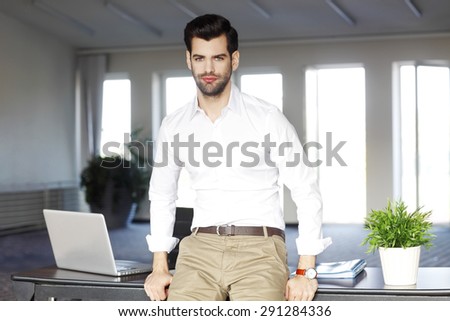 Portrait of young broker standing at office in front of desk.  Confident professional looking at camera and smiling.