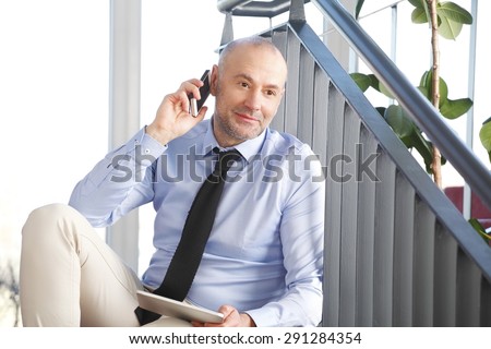Portrait of senior manager sitting at staircase at office building. Busy businessman making call while holding hand digital tablet.