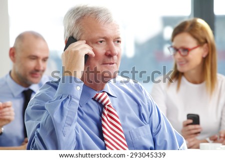 Portrait of senior director sitting at office and talking on cellphone while business people working at background. Business meeting.