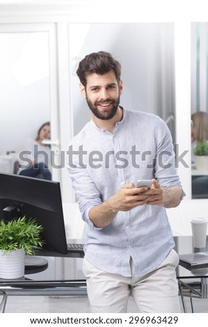 Portrait of young professional man standing at desk in front of computer while holding hand mobile and reading news.