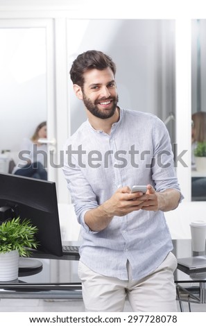 Portrait of young professional man standing at desk in front of computer while holding hand mobile and writing email.