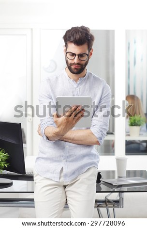 Portrait of young professional man standing at desk in front of computer while holding hand digital tablet and surfing on internet.