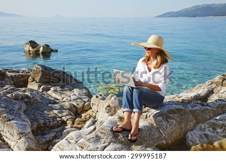 Full length portrait of casual woman working by the sea on her vacation. Middle age female sitting on the rock and typing on her laptop.