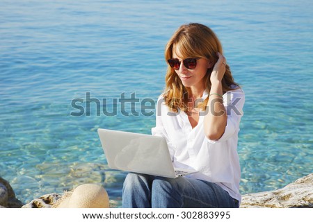 Portrait of smiling woman sitting at the rock by the see with her laptop and using private banking.