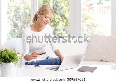 Portrait of smiling woman sitting at sofa in front of laptop and holding credit card in her hand while shopping online.