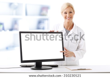 Portrait of smiling pharmacist working on computer while looking at camera and points out white blank monitor.