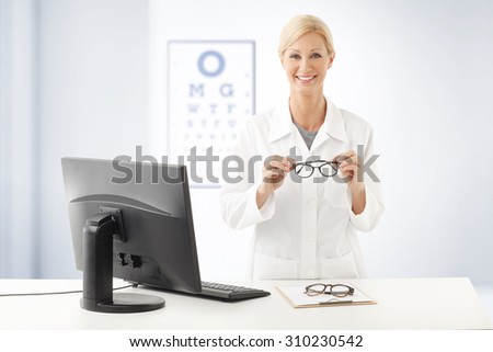 Portrait of smiling optometrist doctor standing ad desk in front of computer and holding hands eyewear. In the background with an eye test chart on the wall.