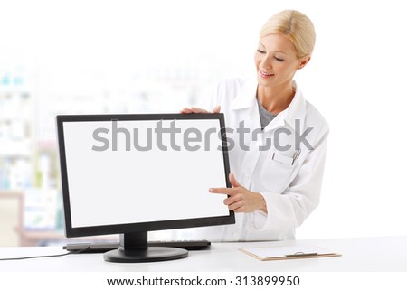 Portrait of smiling pharmacist standing behind the computer and points to the white blank screen.