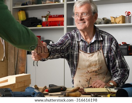 Portrait of retired manual worker sitting at desk in his workshop and shaking hands with casual man.