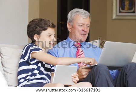 Portrait of young boy teaching her grandfather how to use internet safety. Teenager boy with digital tablet sitting at sofa while senior man using laptop.