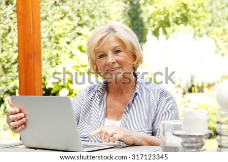 Portrait of senior woman relaxing at home. Retired female typing on computer while sitting in the garden and looking at camera.