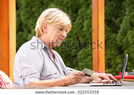 Portrait of senior woman sitting at garden with her laptop. Smiling grandmother holding bank card in her hand while enjoys the convenience of banking online.