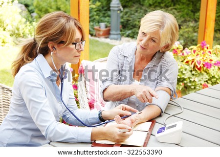 Elderly woman sitting in the garden with home health care nurse an using blood pressure. Home caregiver female consulting with her patient.