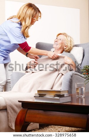 Portrait of smiling nurse cares for elderly woman. Senior female sitting at home in her armchair while home caregiver covering with blanket.