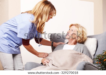 Portrait of smiling nurse cares for elderly woman. Senior female sitting at home in her armchair while home caregiver covering with blanket.