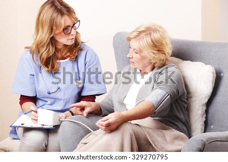 Portrait of senior woman sitting at home and social healthcare nurse checks blood pressure of elderly woman.