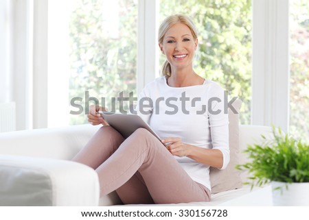 Portrait of cheerful middle age woman sitting at sofa and using her digital tablet while working online at home.