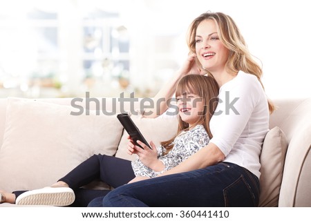 Portrait of blond mother and her cute daughter watching tv together at home. Little kid holding in her hand a remote controll while sitting next to her mom at sofa in living room.
