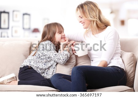 Portrait of a beautiful mother and her little girl sitting at home and sharing a happy moment together. Blond mom holding in her hand a cup of tea.