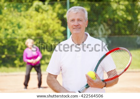Portrait of an active senior man standing at tennis court and holding in hands tennis racket while elderly woman ready to play tennis at background.