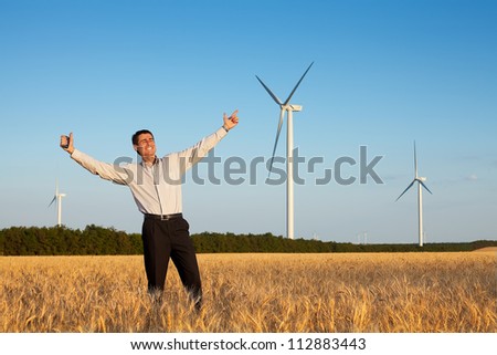 happy farmer (businessman) standing in wheat field over wind turbines background with his hands up and showing ok (thumbs up)