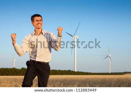 happy farmer (businessman) standing in wheat field over wind turbines background, dancing and holding his hands up
