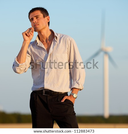 businessman (farmer) standing in a wheat field over background of blue sky and white wind turbine with hands in pockets and wheat spikelet in mouth