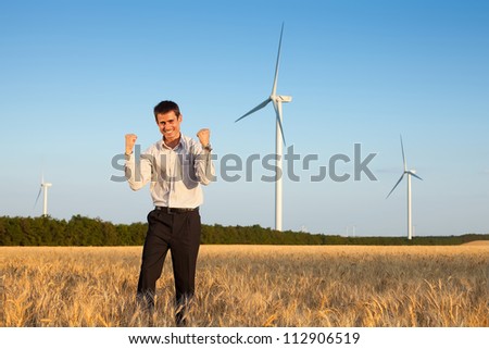 happy farmer (businessman) standing in wheat field over wind turbines background and holding his hands up