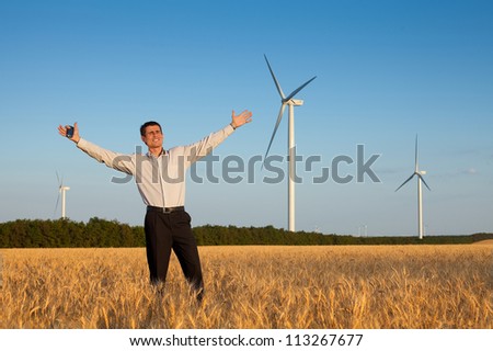 happy businessman (farmer) standing in a wheat field and smiling over background of blue sky and white wind turbine with hands raised