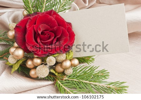 Christmas decoration and blank card. Red rose with golden berries and twigs of a Christmas tree and a blank card for a festive message. On a golden tablecloth.