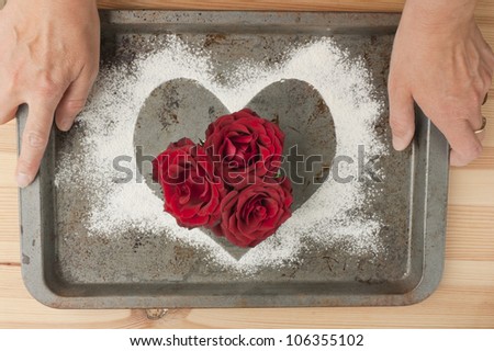 Baking, cooking, taking care of the loved ones. Healthy heart diet concept. Middle aged woman\'s hands holding old grunge metal baking tray with a bunch of red roses and a heart shape made of flour.