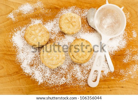 Christmas baking. Home made mince pies on a scratched wooden table covered with icing sugar, next to sieve and metal teaspoon. Snow flake shape pattern on mince pies.