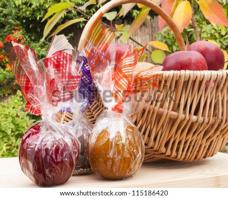 Caramel, chocolate and toffee apples wrapped up in colorful foil next to a basket of freshly picked up red apples. On a wooden table in the garden, against autumn background. Natural light.