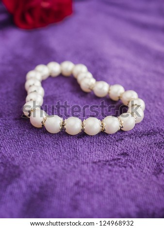 Pearl bracelet in a shape of a heart on purple velvet textile with a single red rose in the background. Concept for a Valentine\'s Day, anniversary or birthday gift. Shallow depth of field.
